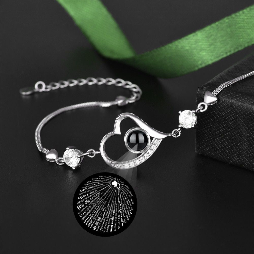Silver bracelet with heart-shaped clasp and engraved disc on a dark surface with green ribbon.