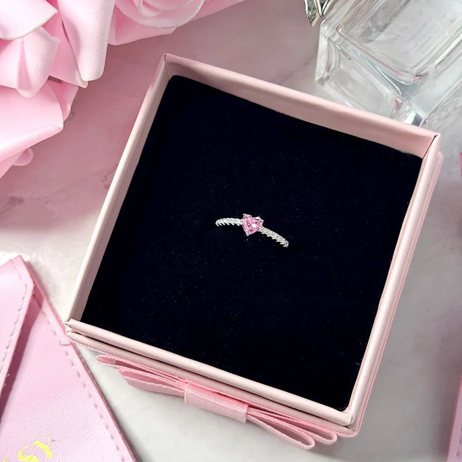 Pink ring on a black cushion in a pink jewelry box with pink flowers and a glass container in the background.