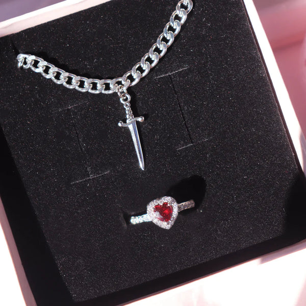 A jewelry set presented in a box, featuring a heart-shaped ring and a sword-shaped bracelet, both with silver-colored bands.