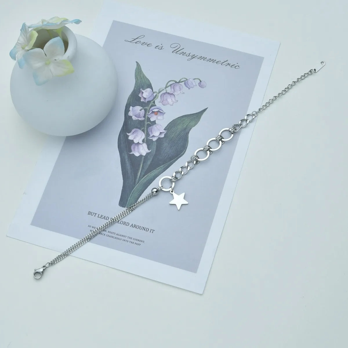 Silver bracelet with star charm on a card with floral design and text, next to a small vase with flowers.