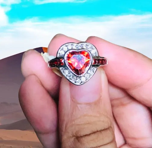 Heart-shaped ring with a red gemstone held between fingers against a blurred natural background