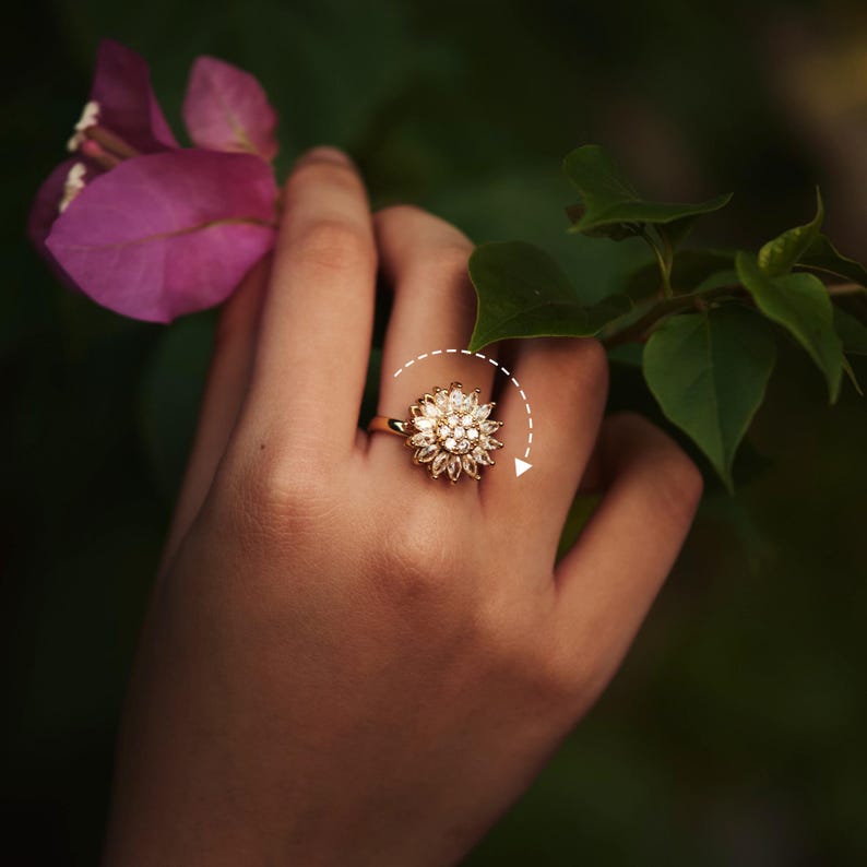 Hand wearing a gold ring with a floral design, surrounded by green leaves and a pink flower.